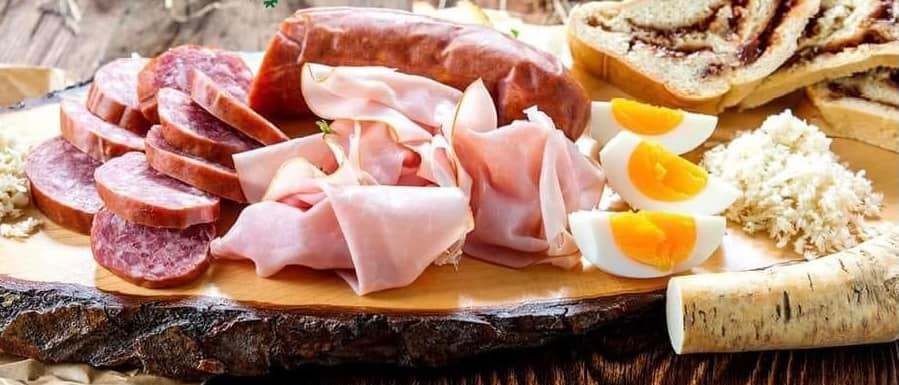 Ein Holztablett mit verschiedenen Fleischsorten, darunter Scheiben von Salami, Schinken und Eiern. Daneben befindet sich auch ein Stück Zimtbrot.