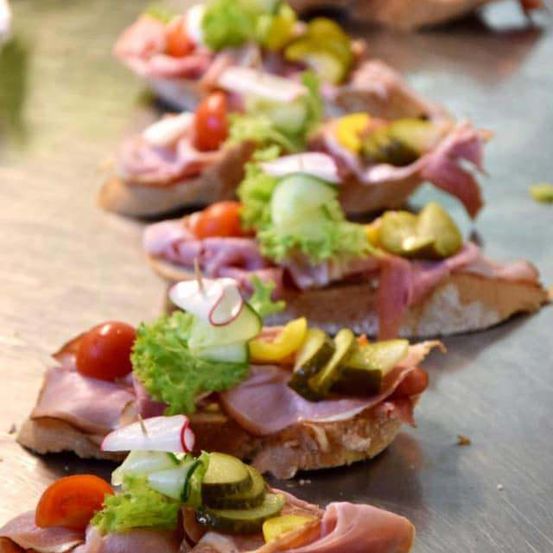 Eine Nahaufnahme von mehreren Scheiben getoastetem Brot, belegt mit Schinken, Tomaten, Gurken und Salat, auf einer Metalloberfläche angeordnet.