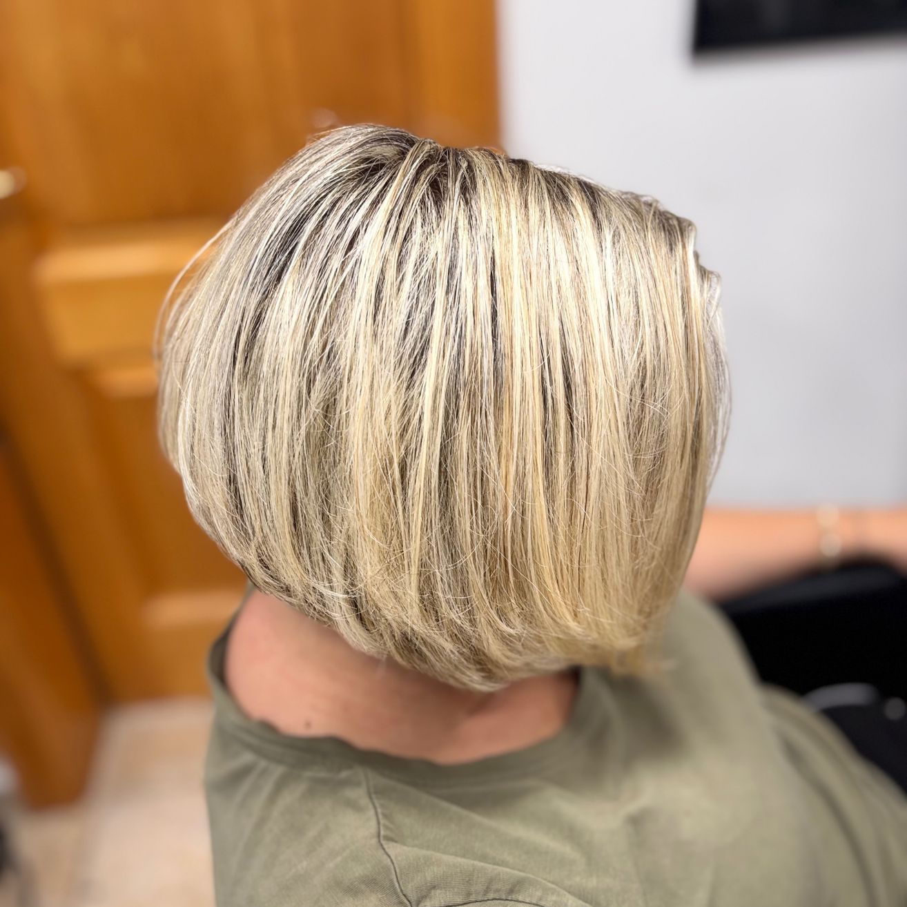Eine Frau mit kurzen, blonden Haaren in einem grünen T-Shirt sitzt in einem Salon. Sie blickt von der Kamera weg, mit einer hölzernen Tür und einer weißen Wand im Hintergrund.