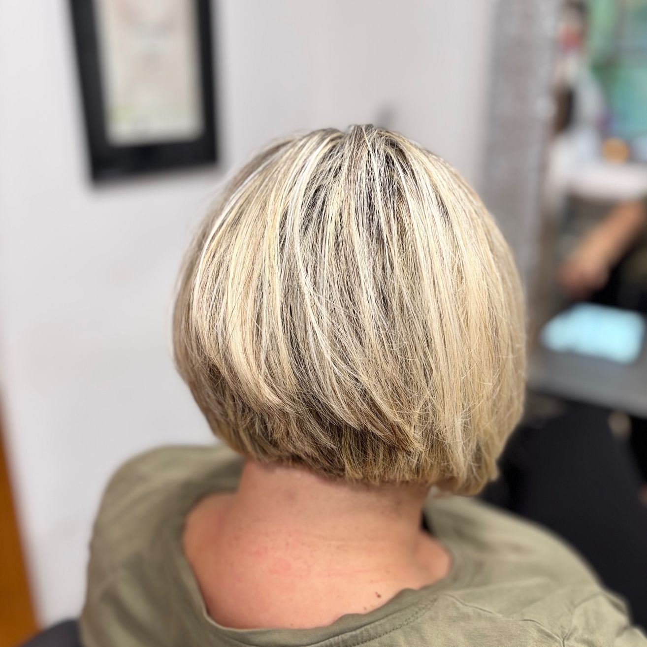 Eine Frau mit kurzen blonden Haaren sitzt in einem Salon und trägt ein grünes Shirt. Der Hinterkopf ist sichtbar und zeigt einen Haarschnitt. Ein gerahmtes Bild hängt an der Wand hinter ihr.