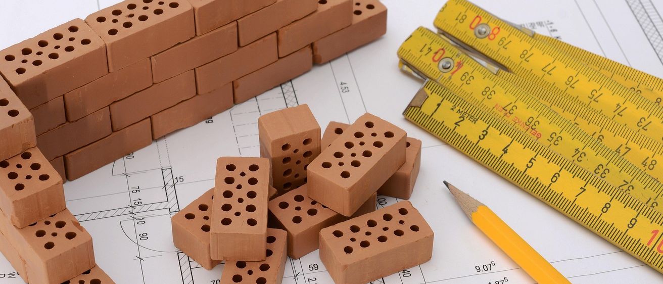 Ein Stapel Lehmziegel und ein gelbes Maßband liegen auf einem Bauplan mit Zahlen und Linien. Ein Bleistift ist ebenfalls auf dem Bauplan abgelegt.