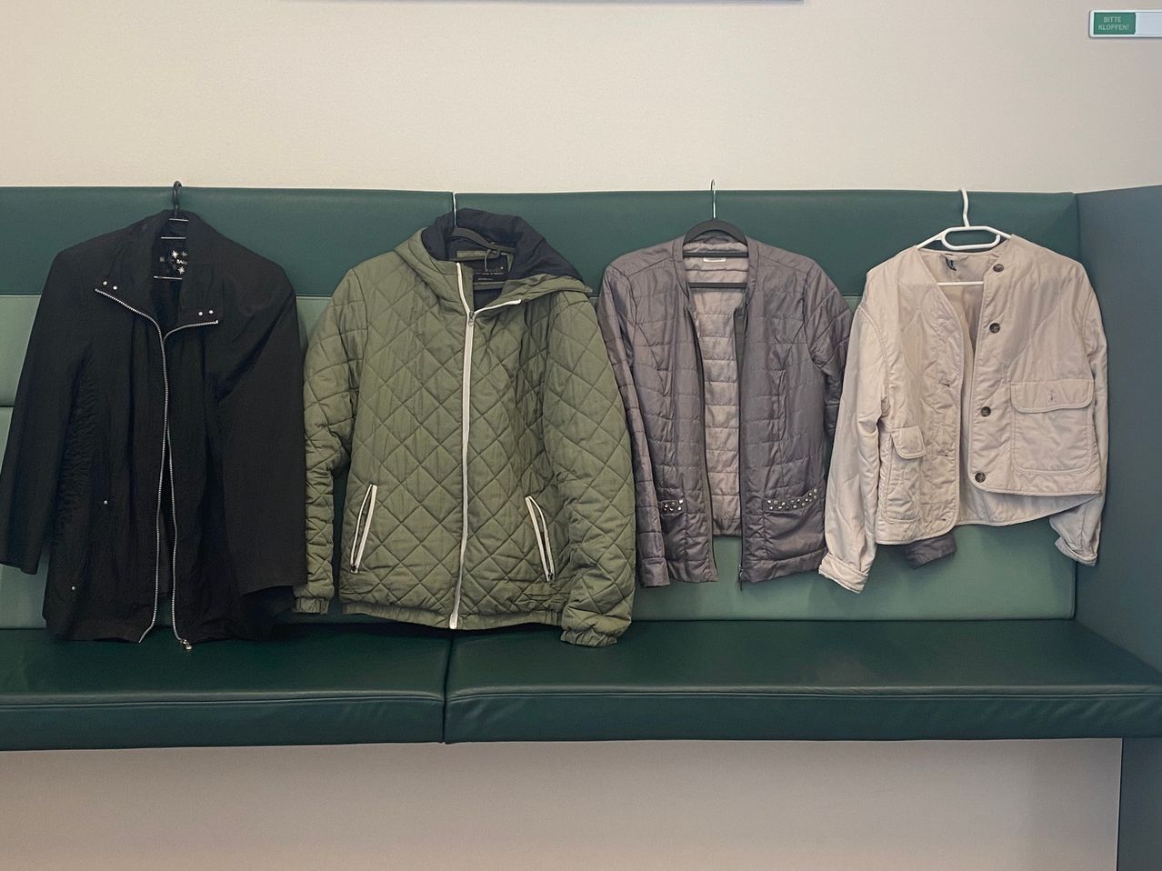 Five jackets in different colors and styles hang on a green bench against a white wall. The jackets are black, green, gray, and beige.