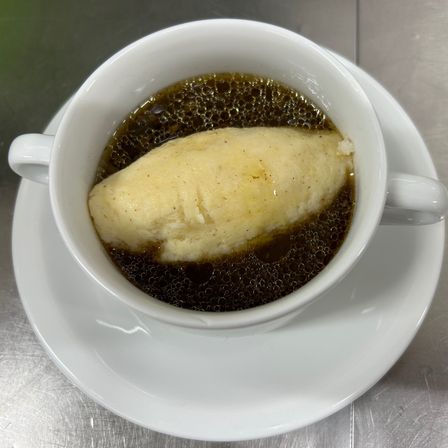 Eine Tasse mit einem cremigen weißen Pudding, der in einer braunen Flüssigkeit auf einem weißen Unterteller auf einer metallischen Oberfläche schwimmt.