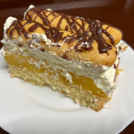 Ein Stück Kuchen mit Sahne und Schokoladenglasur auf einem weißen Teller. Es hat Schichten aus Kuchen, Sahne und einer gelben Füllung. Zwei Gebäckstäbchen sind obenauf.