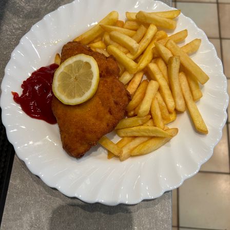 Ein Teller mit gebratenem Schnitzel, Pommes Frites und Ketchup mit einer Zitronenscheibe obenauf, auf einer gefliesten Oberfläche.