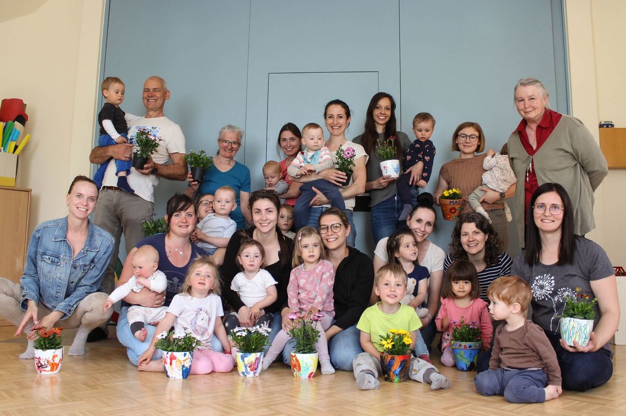 A group of adults and children pose for a photo with flowers in pots, all smiling in front of a light blue wall.