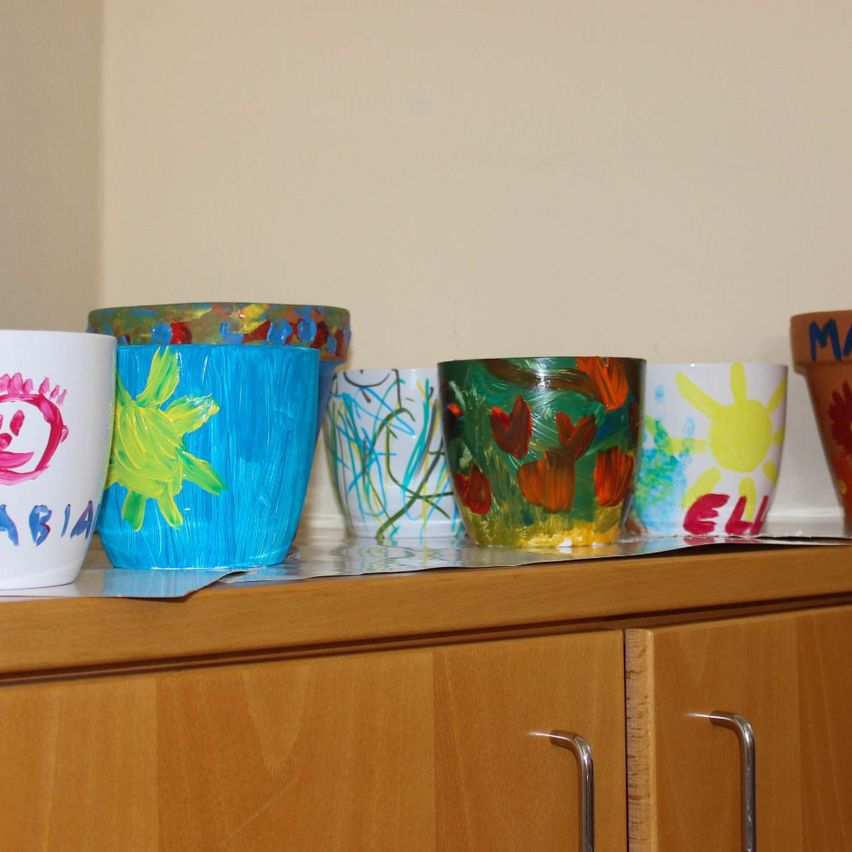 Multiple colorful pots, each with unique designs, are arranged on a wooden shelf. Each pot has a name written on it, such as 'MARIA', 'ELIA', and 'ABIA'.
