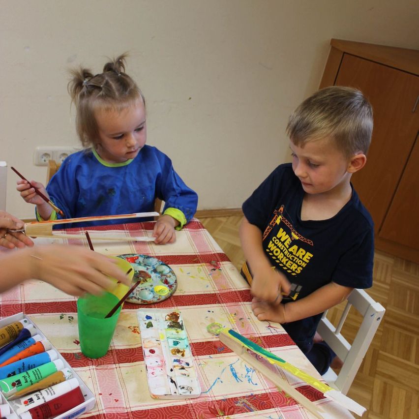 Two children are sitting at a table, engaged in a painting activity. A girl is using a paintbrush while a boy watches. Various painting tools and colors are on the table.