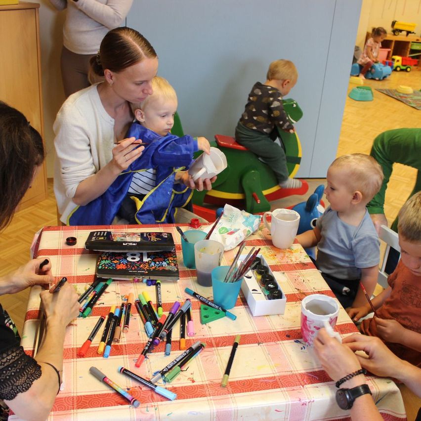 A group of young children and adults are in a room. One woman holds a baby, and others are painting. A table has pens, cups, and a book. Other children play with toys.