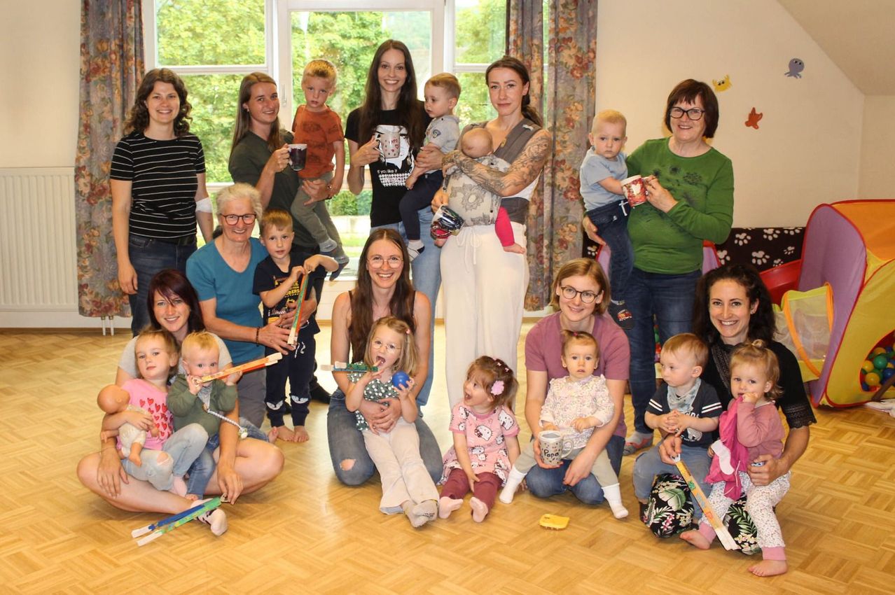 A group of adults and children are gathered indoors, some standing and others sitting on the floor. They hold cups, toys, and babies, smiling and posing for a photo.