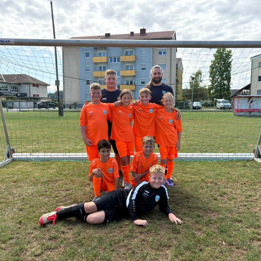 Eine Fußballmannschaft posiert für ein Gruppenfoto vor einem Tor, im Hintergrund Gebäude und Bäume.
