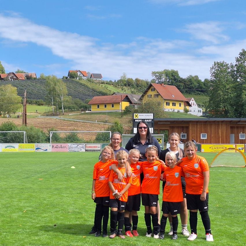 Eine Fußballmannschaft junger Mädchen in orangefarbenen Trikots steht auf einem grünen Feld mit einer Frau in Sonnenbrille hinter ihnen. Hinter dem Feld stehen Häuser und ein Schild. Der Himmel ist klar und blau.