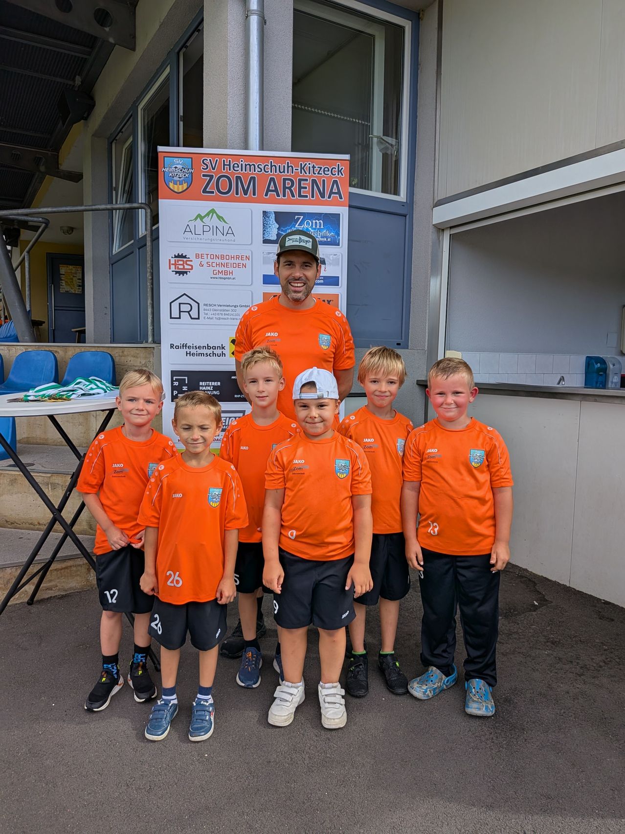 Ein Mann in einem orangefarbenen Shirt steht hinter einer Gruppe Jungen in orangefarbenen Uniformen. Sie stehen vor einem Gebäude mit einem Banner, das SV Heimschuh-Kitzeck ZOM ARENA beschriftet ist.