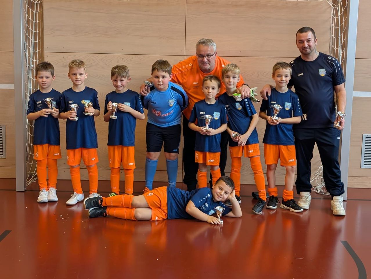 Eine Gruppe junger Jungen in blauen und orangefarbenen Trikots posiert für ein Foto mit ihren Trophäen. Zwei Erwachsene stehen hinter ihnen, einer hält eine Trophäe. Sie alle lächeln und stehen in einem Raum.
