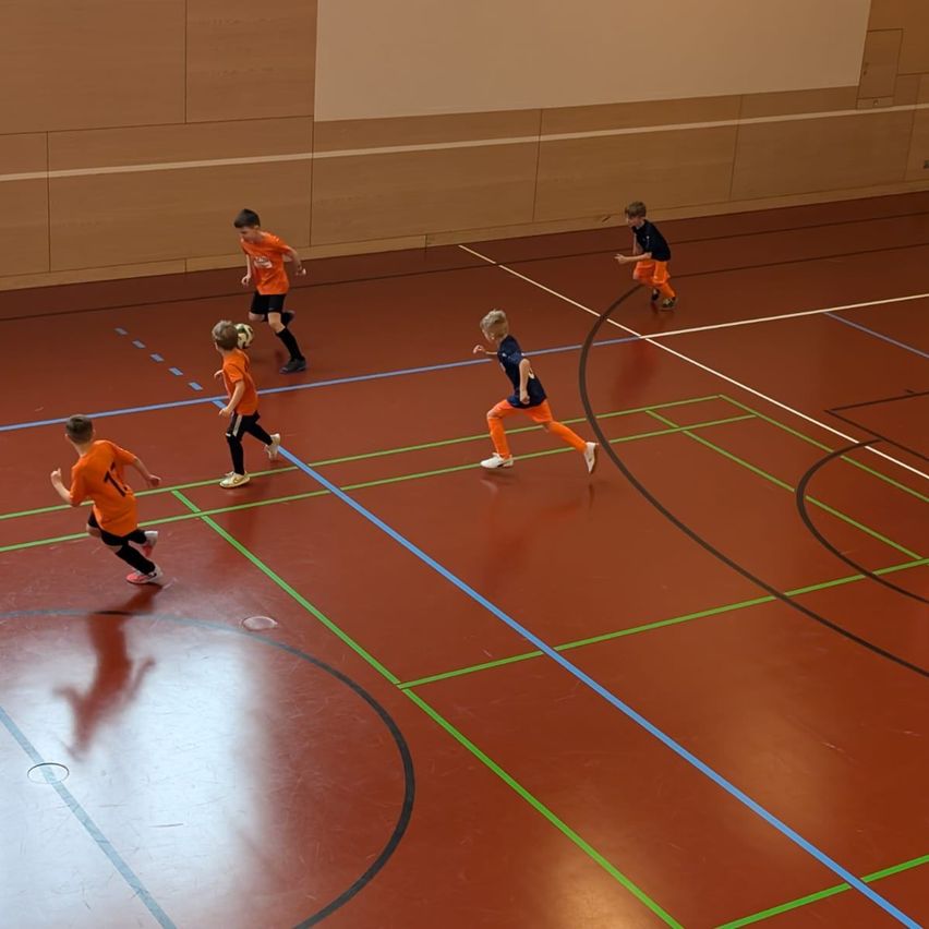 Eine Gruppe von Kindern spielt Fußball auf einem Hallenplatz. Sie tragen orangefarbene und blaue Trikots. Der Platz hat blaue und grüne Markierungen.
