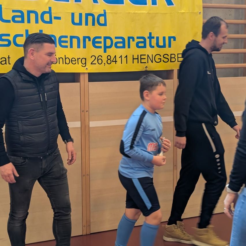 Drei Personen gehen in der Nähe eines Banners, das 'Land- und Baumaschinenreparatur' bewirbt. Der Mann in der Mitte trägt ein blaues Shirt, während die anderen schwarze Jacken tragen.