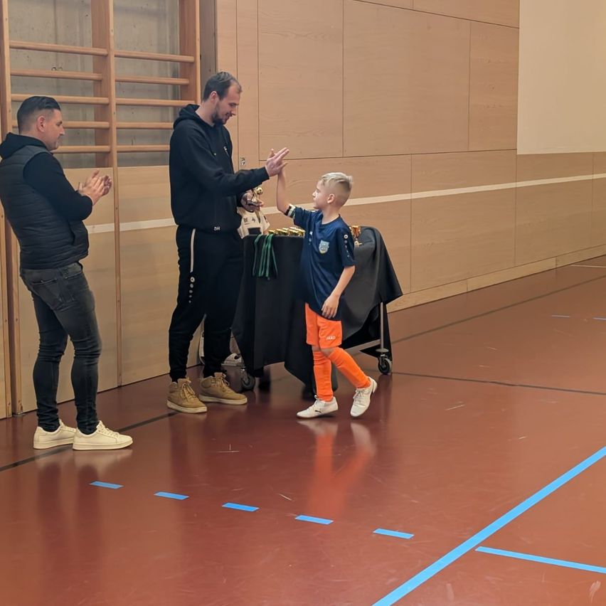 Ein junger Junge in orangefarbenen Hosen und Turnschuhen erhält von einem Mann einen Pokal, während ein anderer Mann in einer Turnhalle mit blauen Linien auf dem Boden klatscht.