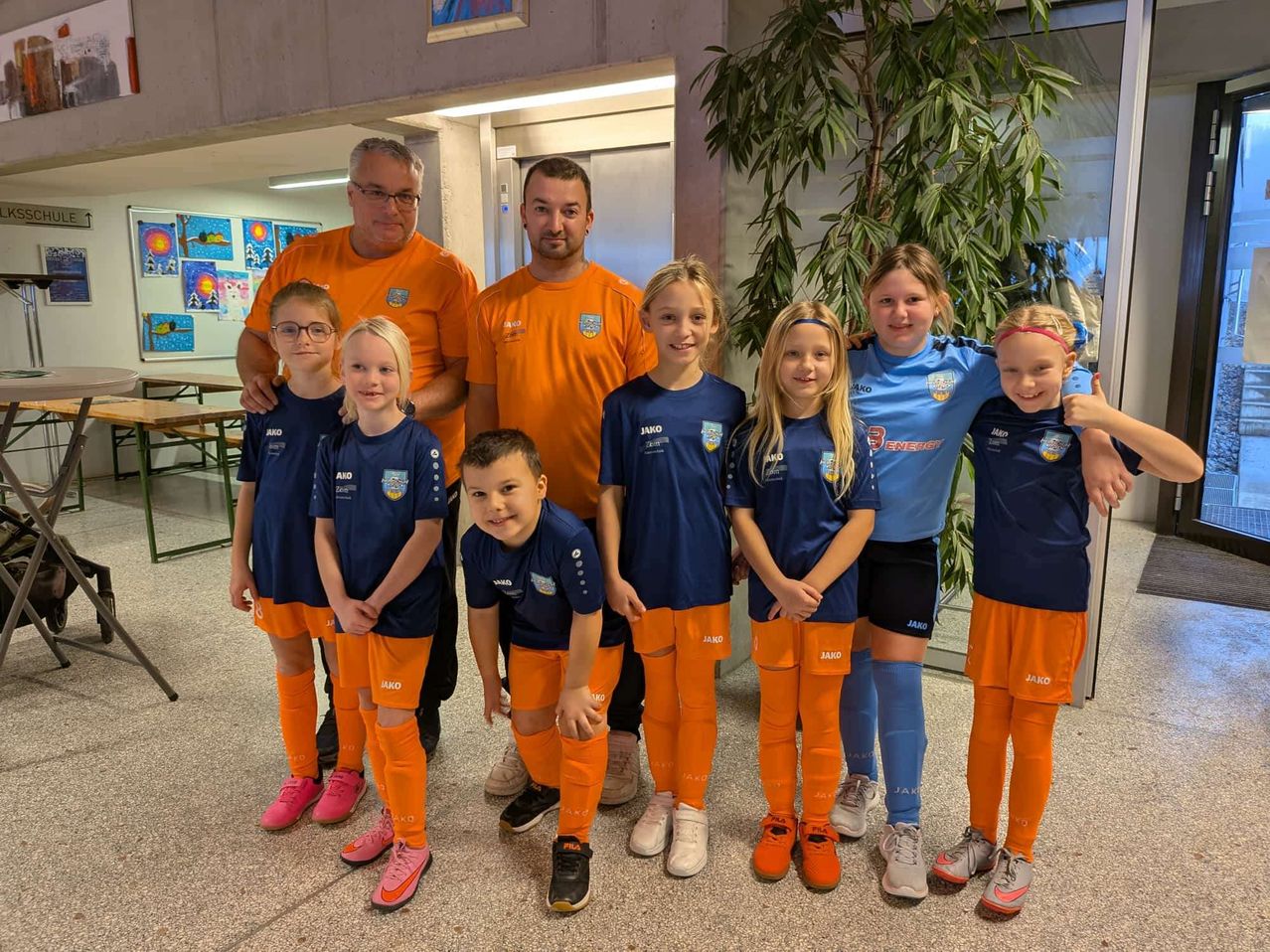 Eine Gruppe junger Fußballspieler in blauen und orangefarbenen Trikots posiert für ein Foto mit ihren Trainern.