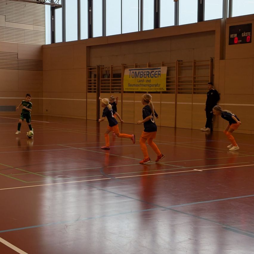 Eine Gruppe von Kindern spielt Hallenfußball. Sie tragen orangefarbene Uniformen und Turnschuhe. Ein Junge in Grün tritt den Ball. Zwei Erwachsene beobachten von der Seitenlinie.