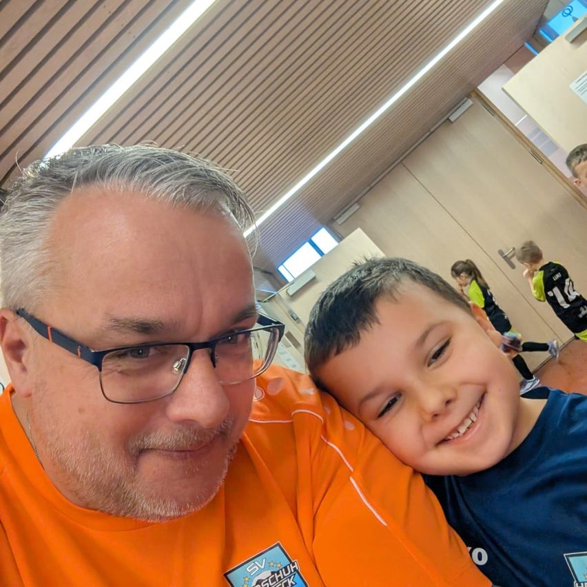 Ein erwachsener Mann mit grauem Haar und Bart trägt ein orangefarbenes T-Shirt. Er posiert für ein Foto mit einem lächelnden jungen Jungen in einem blauen T-Shirt. Sie sind in einer Sporthalle.