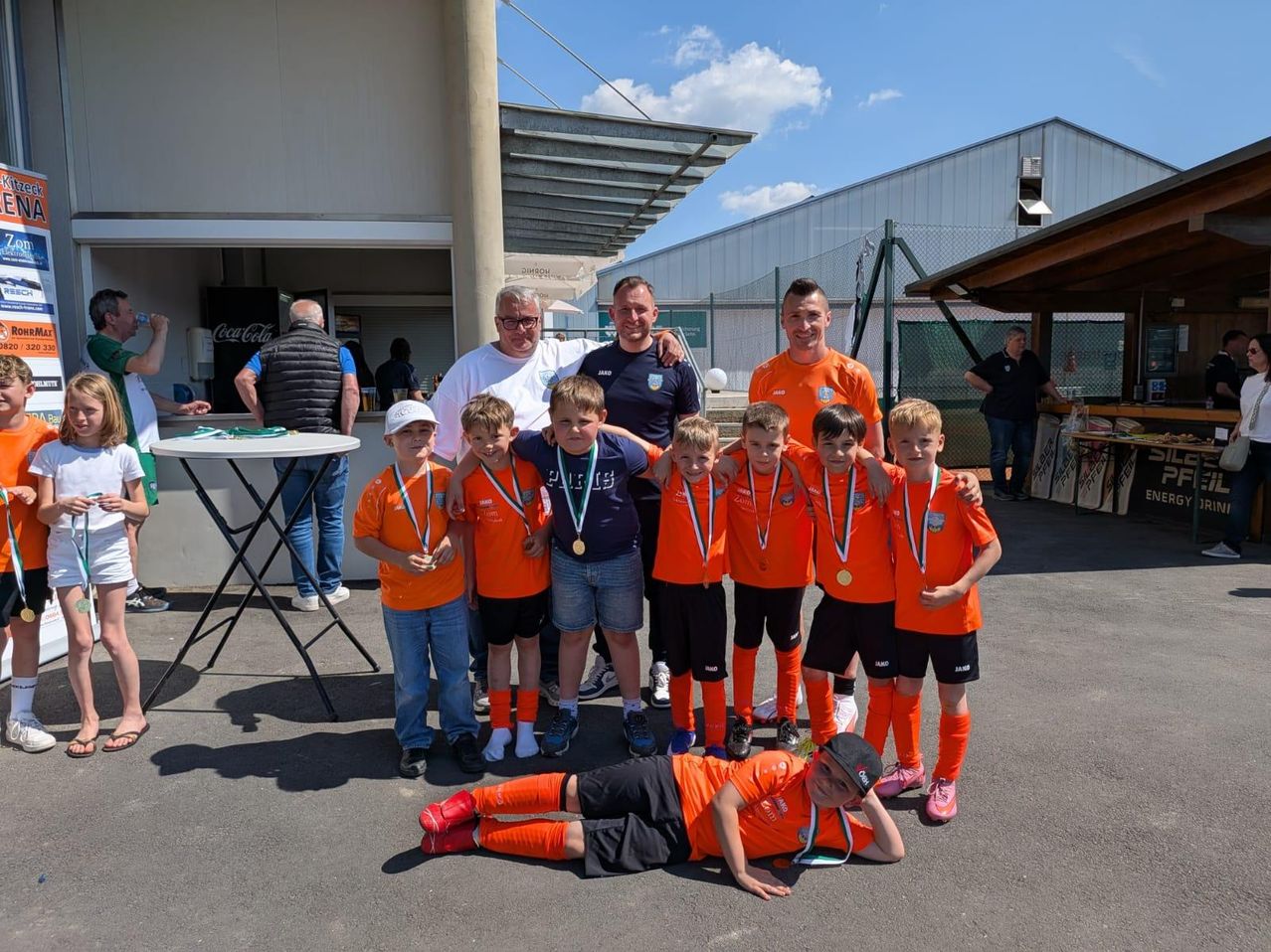 Eine Gruppe junger Fußballspieler, gekleidet in orangefarbenen Trikots, posiert für ein Foto mit ihren Trainern. Sie halten ihre Medaillen und ein Spieler liegt auf dem Boden. Im Hintergrund befindet sich ein Gebäude mit einem Coca-Cola-Schild.