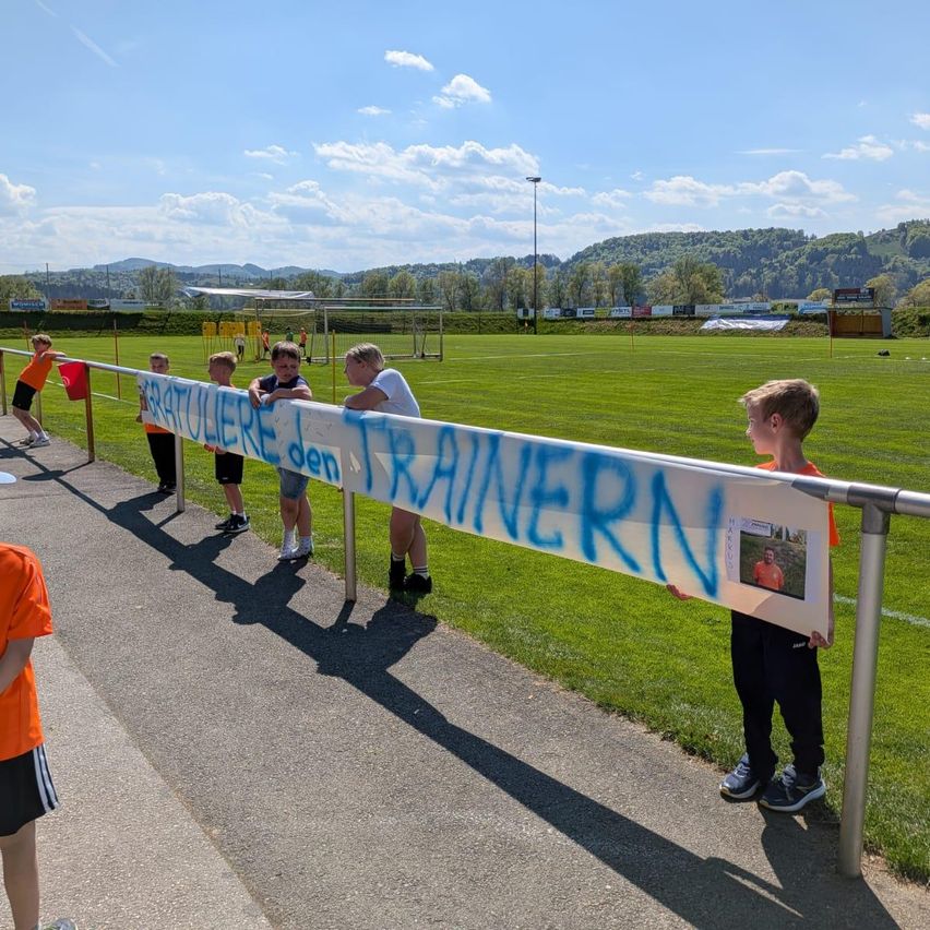Mehrere Kinder stehen auf einem Bürgersteig und halten ein Banner mit einem Foto und Text. Sie befinden sich auf einem Fußballfeld.