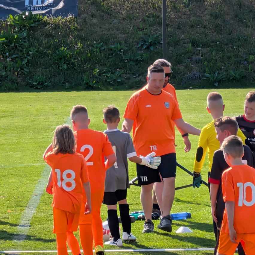 Fußballspieler in orangefarbenen Trikots, einschließlich eines Trainers, versammeln sich auf einem Feld. Sie stehen in der Nähe einer weißen Linie mit einem Ball auf dem Boden.