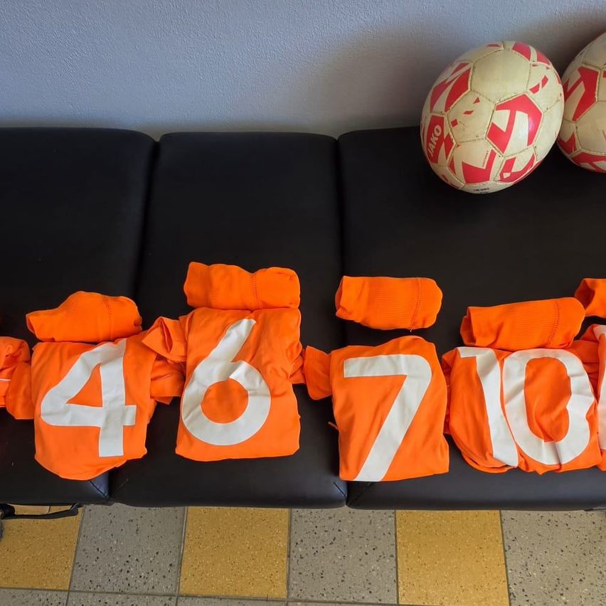 Orangefarbene Trikots mit den Nummern 4, 6, 7 und 10 auf einem schwarzen Sofa, mit zwei Fußballbällen im Hintergrund.