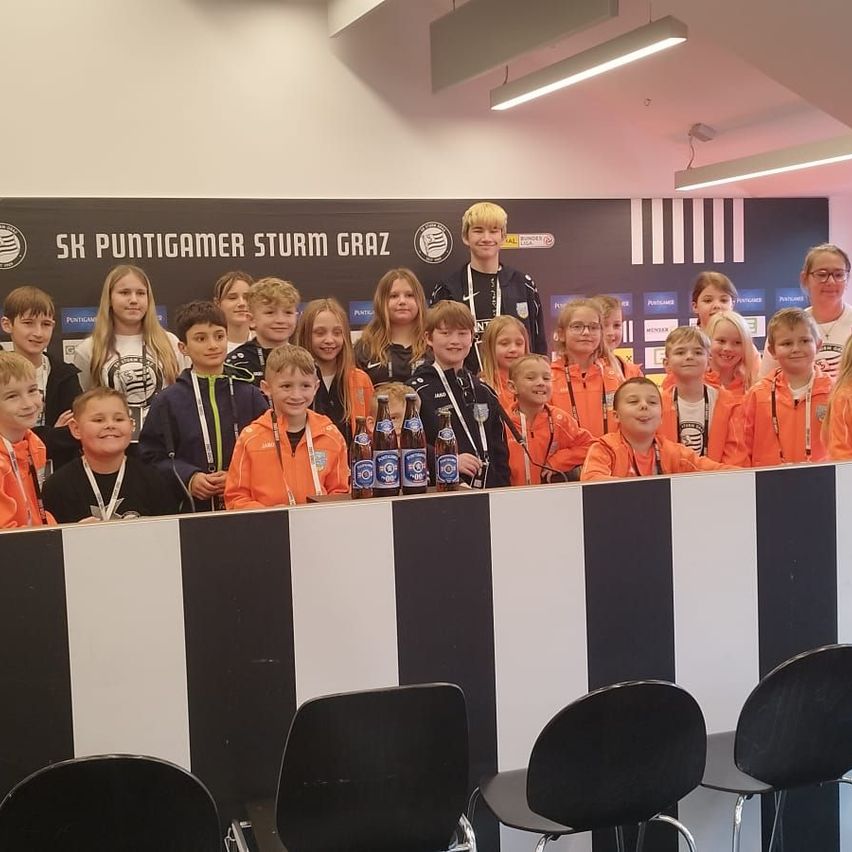 A group of children and a few adults are posing for a photo, likely in a competition. They wear orange jackets, some with name tags. Behind them, a banner reads 'SH PUNTIGAMER STURM GRAZ.' Bottles are on the desk.