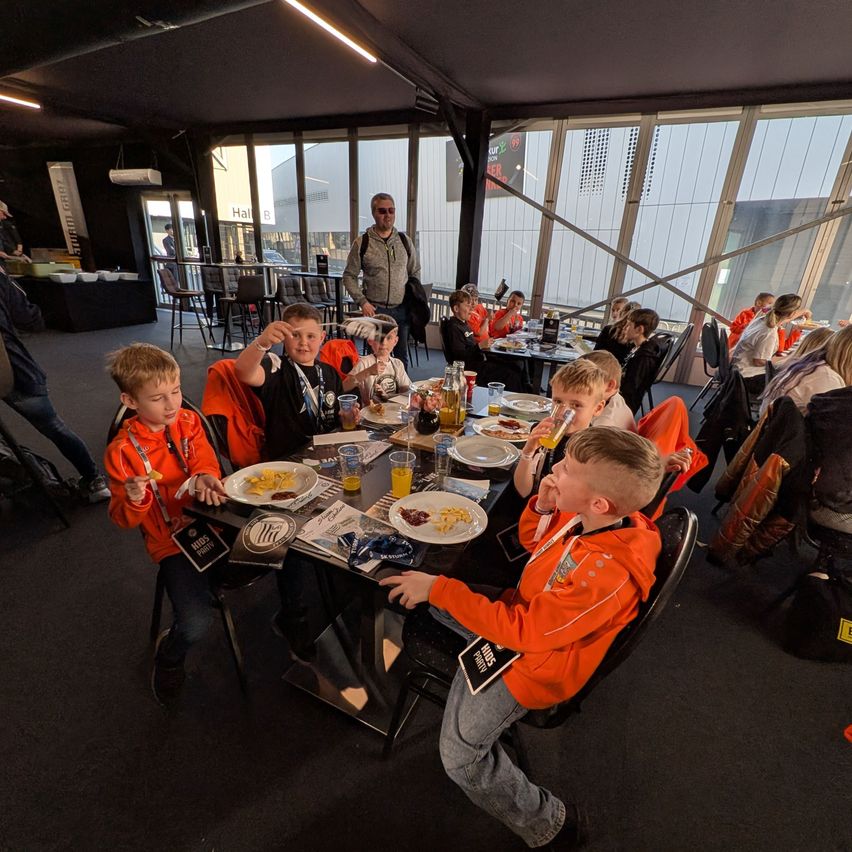 Several children in orange jackets sit around a table eating. A man stands near the table, while others sit at separate tables. The room has glass walls and ceilings with a view of the outside.