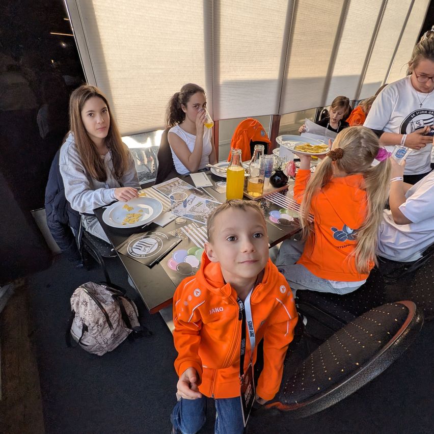 A group of people are seated around a table in a restaurant. A young boy in an orange jacket stands in front of the table. The table is set with plates, bottles, and other items. Some people are eating and drinking. A backpack is on the floor.