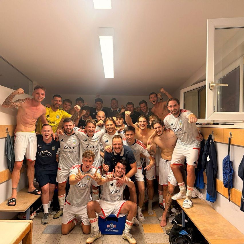 A group of soccer players are celebrating in a locker room. They are wearing white uniforms with red stripes. Some are shirtless, and they are posing for a photo.