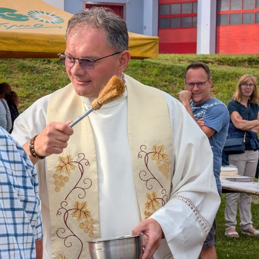 Ein Priester hält eine silberne Schüssel und einen Pinsel in der Hand, trägt eine Brille und ein weißes Gewand mit goldenen Blattdesigns. Hinter ihm stehen eine Gruppe von Menschen, die zuschauen. Ein Gebäude mit roten Türen und ein gelbes Zelt sind im Hintergrund.