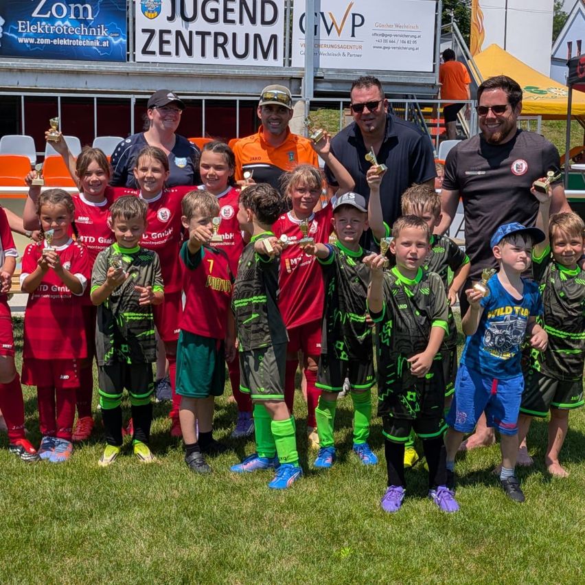 Eine Gruppe junger Fußballspieler in roten und schwarzen Trikots hält auf einem Feld Trophäen. Hinter ihnen stehen zwei Männer und zwei Frauen. Hinter der Gruppe steht ein Schild mit der Aufschrift Jugend Zentrum.