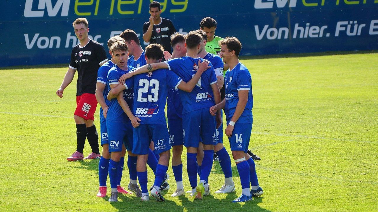 Fußballspieler in blauen Trikots versammeln sich auf einem Feld, die Nummern 23 und 14 sichtbar. Dahinter ein Mann in Schwarz und ein Trainer in Grün. Werbung auf der Umzäunung.