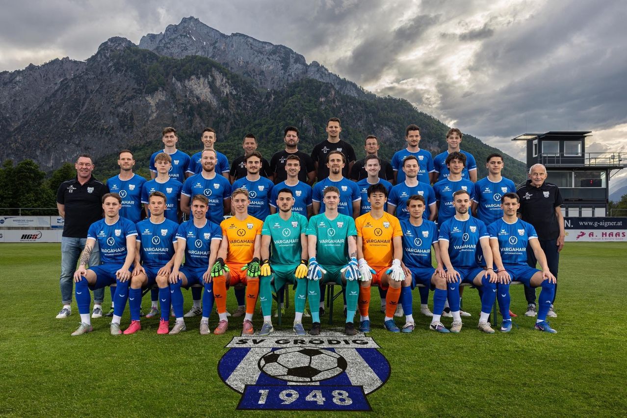 A soccer team poses for a photo in front of mountains. The team wears blue and orange uniforms. They sit on the ground, and a soccer logo is in front of them.