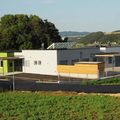 Ein modernes Gebäude mit grüner und weißer Außenfassade befindet sich in einer ländlichen Landschaft. Das Gebäude hat Solarpanele auf dem Dach und ein großes grünes Feld davor. Bäume und Hügel sind im Hintergrund sichtbar.