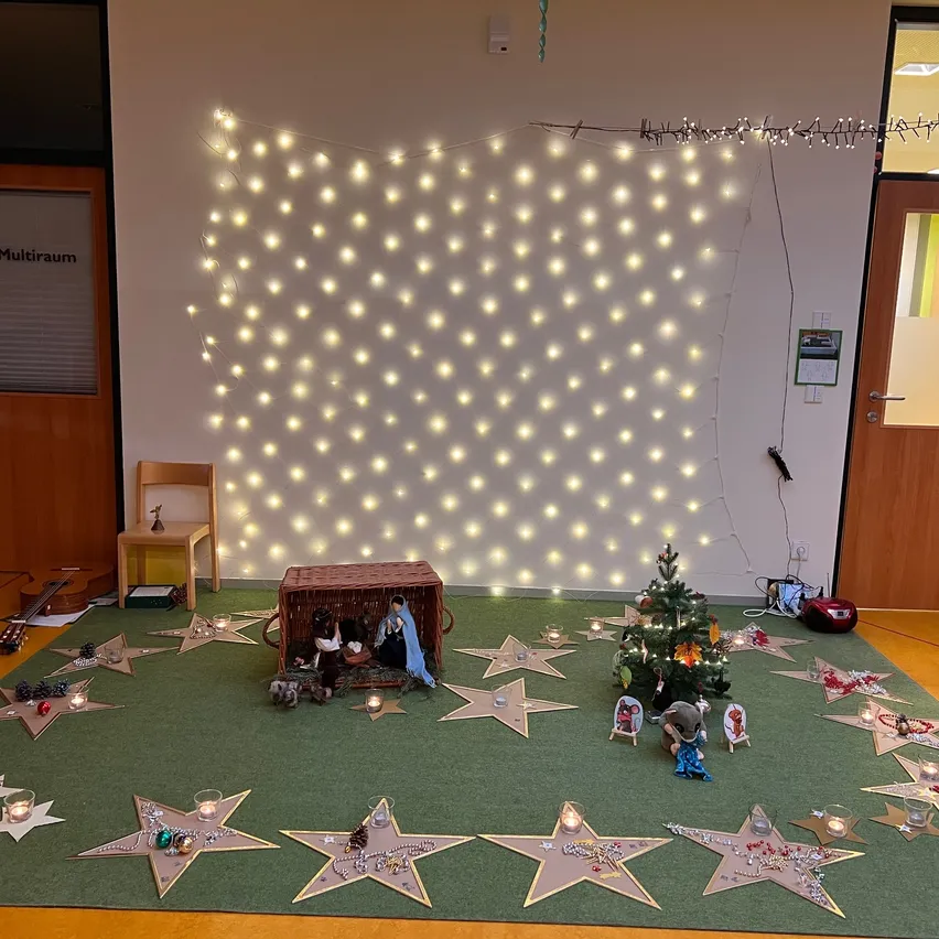 Ein Raum, der für Weihnachten geschmückt ist, mit einer Krippe, einem Weihnachtsbaum und Kerzen. Sterne sind auf dem Boden verstreut. Lichter sind an der Wand und ein Stuhl steht in der Nähe der Tür.