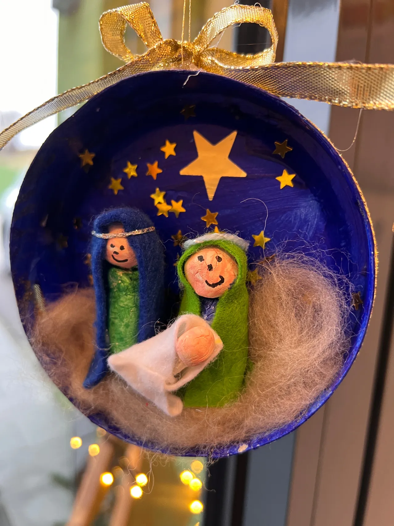 Ein blauer runder Schmuck mit einer Krippenszene, bestehend aus Maria, Josef und Baby Jesus, umgeben von Wolle und Sternen auf blauem Hintergrund.