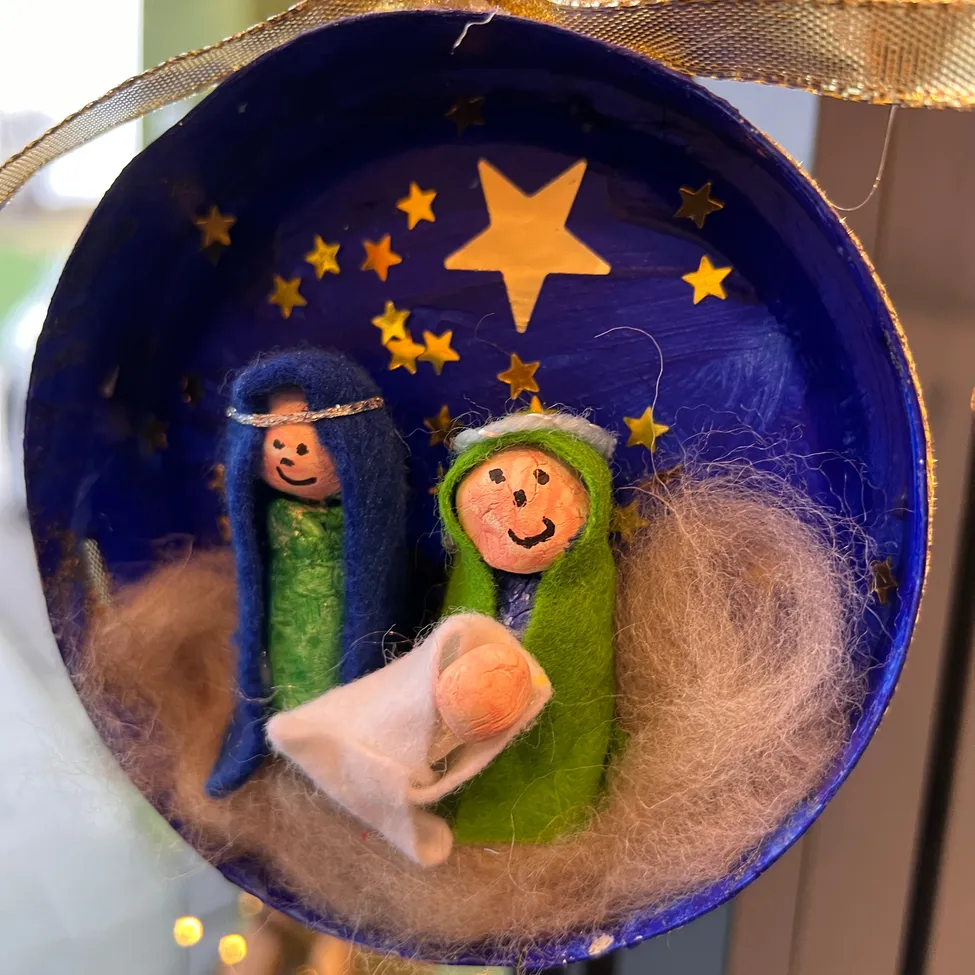 Ein blauer runder Schmuck mit einer Krippenszene, bestehend aus Maria, Josef und Baby Jesus, umgeben von Wolle und Sternen auf blauem Hintergrund.