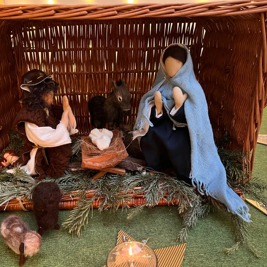 Eine Krippe in einem Weidenkorb, mit Josef, Maria und dem Jesuskind. Die Szene zeigt einen Esel und ein Schaf, umgeben von einer Kerze und Grünpflanzen.