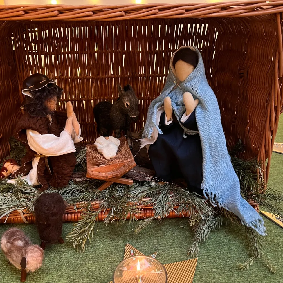 Eine Krippe in einem Weidenkorb, mit Josef, Maria und dem Jesuskind. Die Szene zeigt einen Esel und ein Schaf, umgeben von einer Kerze und Grünpflanzen.