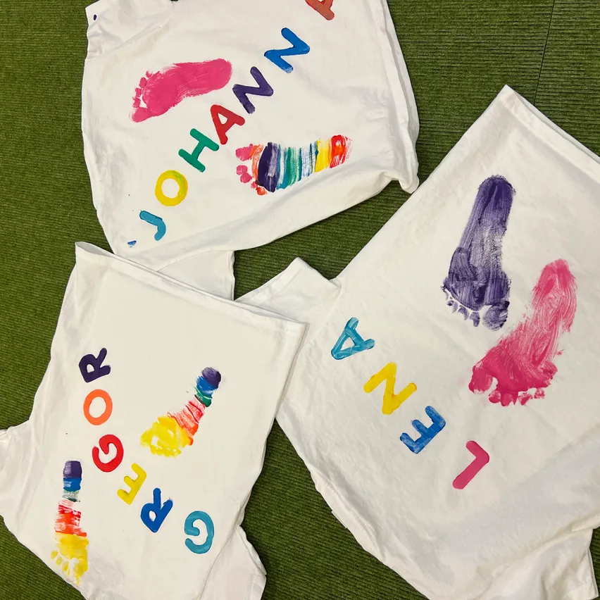 Vier weiße T-Shirts mit bunten Buchstaben und Fußabdrücken. 'JOHANN' oben links, 'FARBE' unten links, 'ZAEGE' unten rechts und 'JAEL' oben rechts.