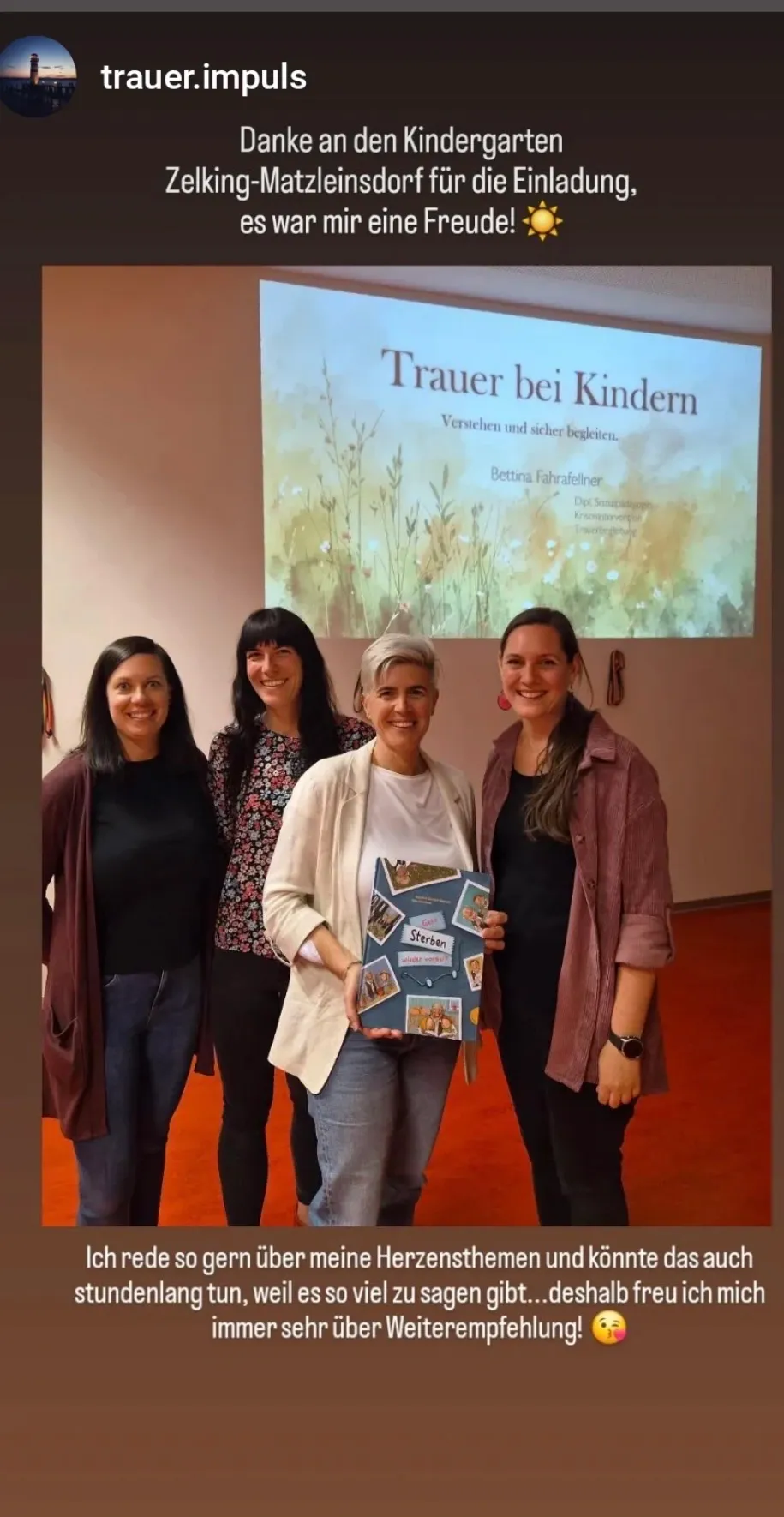 Vier Frauen stehen nebeneinander und lächeln, eine hält ein Buch mit dem Titel 'Sterben.' Hinter ihnen ist eine Leinwand mit dem Titel 'Trauer bei Kindern.' Sie scheinen bei einer Präsentation zu sein.
