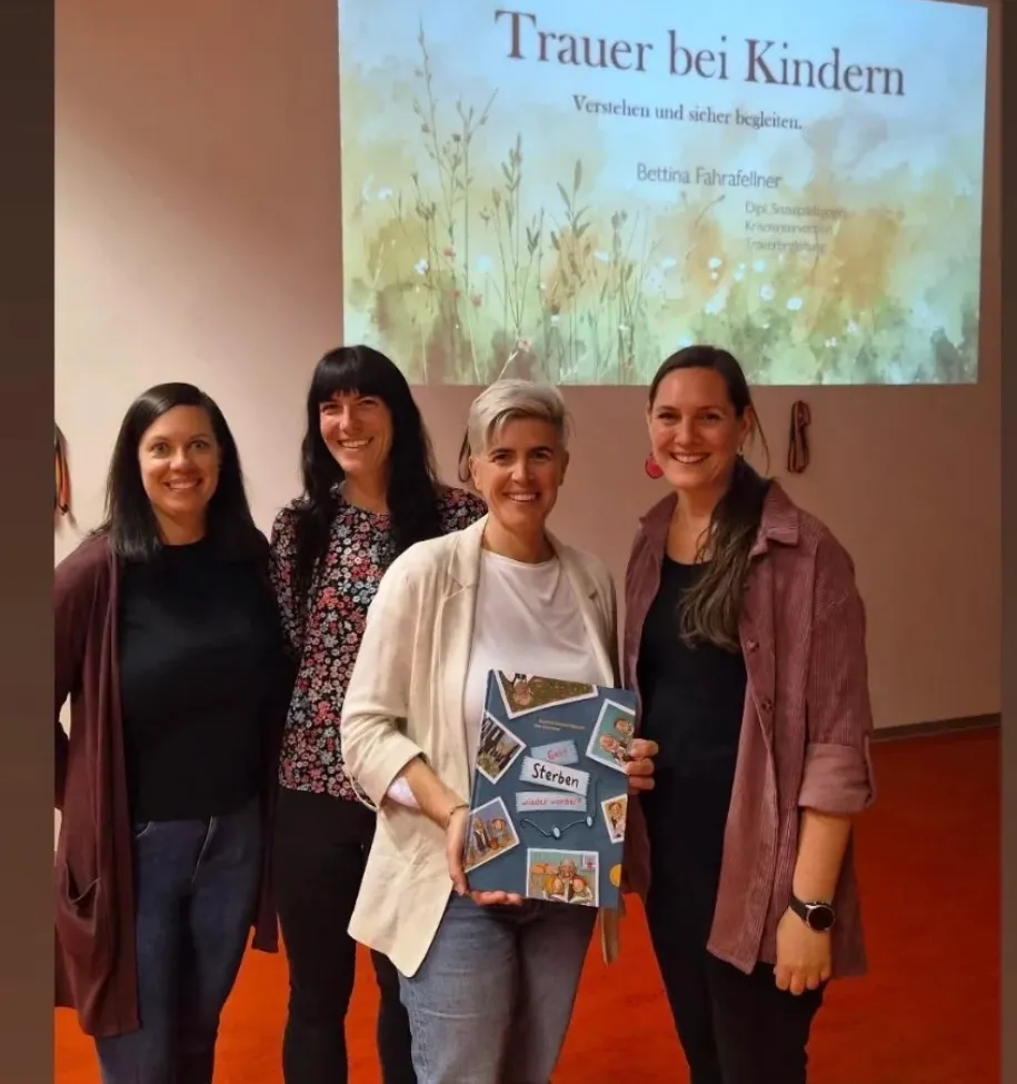 Vier Frauen stehen nebeneinander und lächeln, eine hält ein Buch mit dem Titel 'Sterben.' Hinter ihnen ist eine Leinwand mit dem Titel 'Trauer bei Kindern.' Sie scheinen bei einer Präsentation zu sein.
