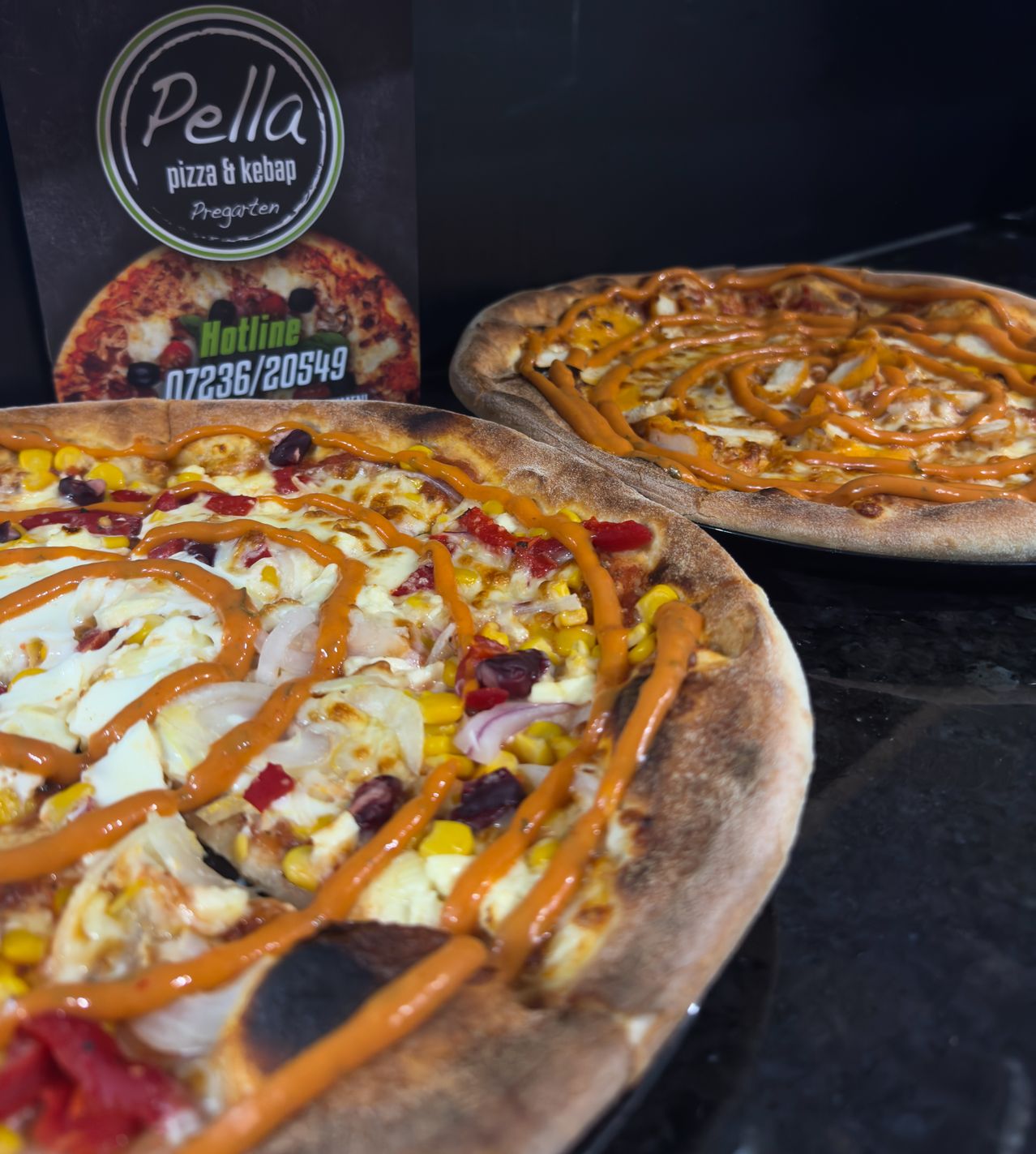 Zwei Pizzen mit verschiedenen Belägen und Soßen auf einer schwarzen Oberfläche. Eine hat Mais, Oliven und Tomaten, während die andere Käse und Pepperoni hat.