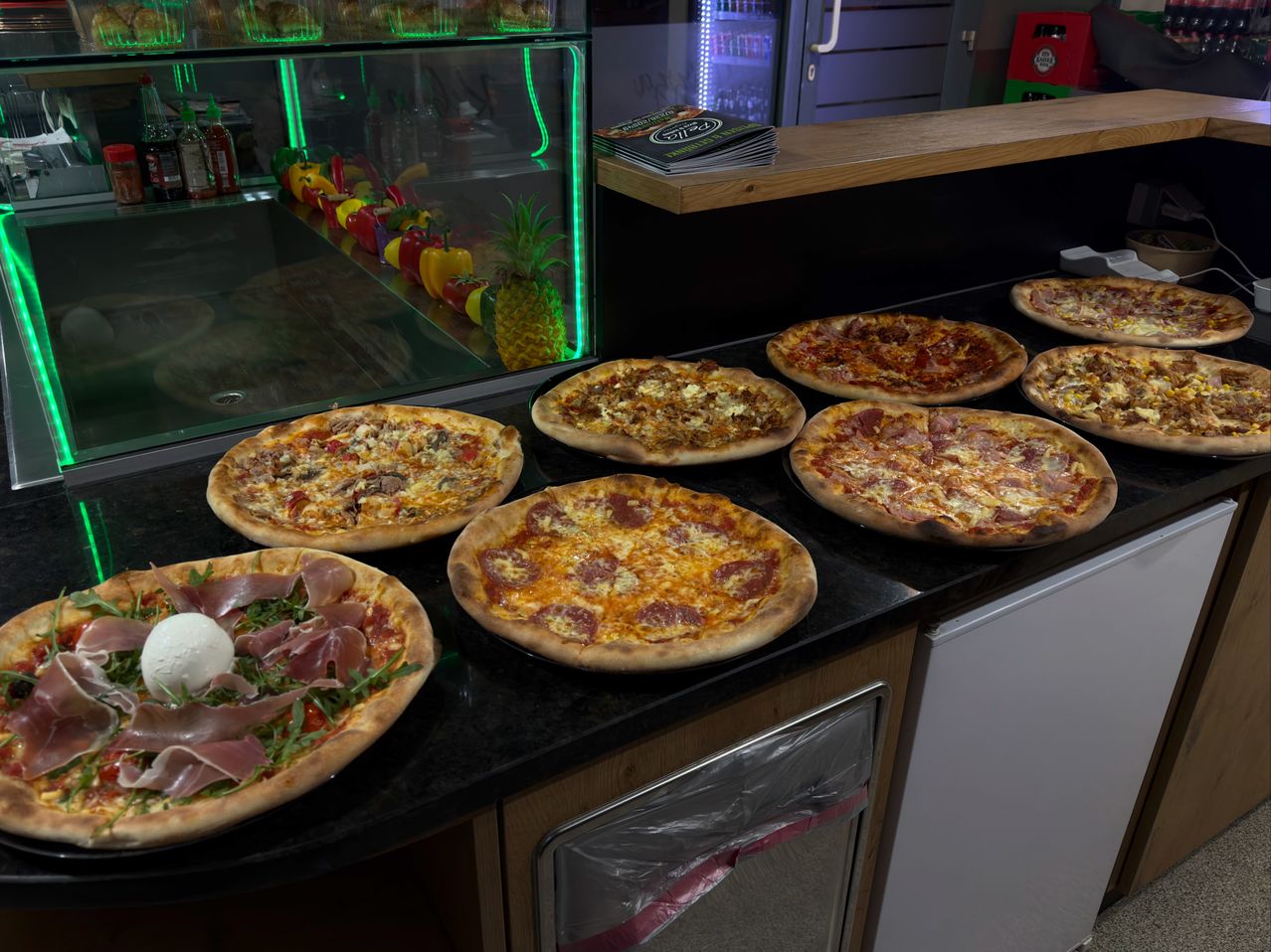 Eine Ausstellung verschiedener Pizzen in einem Restaurant, einschließlich Pepperoni, Käse und Fleischbelägen. Eine Ananas und Flaschen sind auf einem Glasregal. Der Tresen hat hölzerne Oberflächen.
