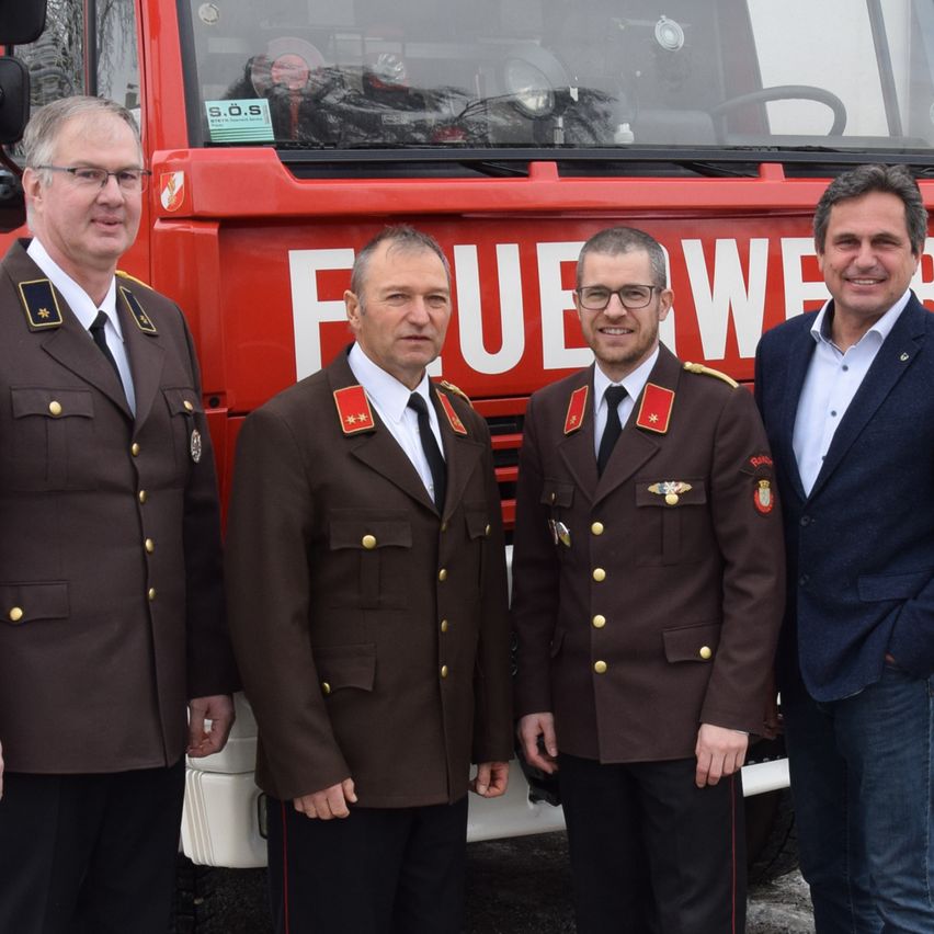 Four men stand in front of a red fire truck with the word 'FEUER' on it. Three men wear military uniforms, and one wears a blue suit. They all look forward and smile.