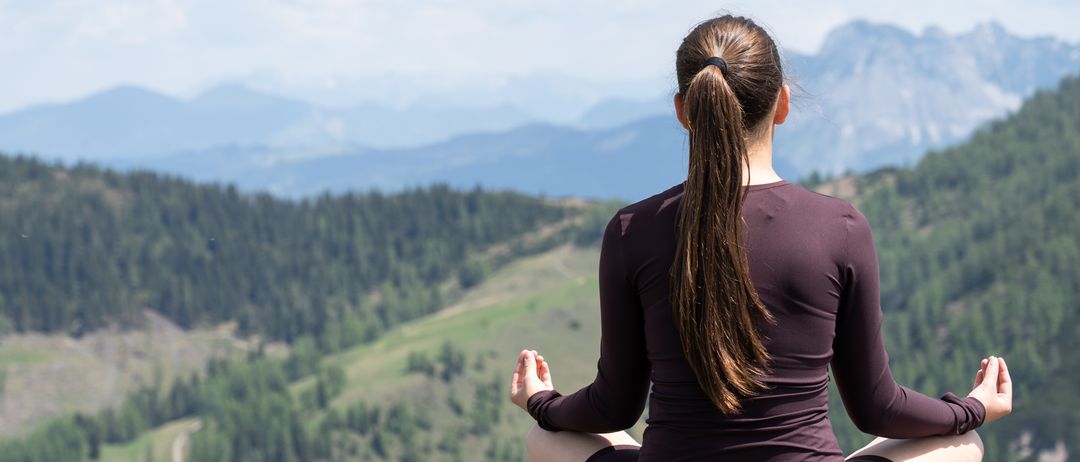 Eine Frau in einem braunen Outfit sitzt mit gekreuzten Beinen auf einem hölzernen Geländer und blickt auf eine Berglandschaft mit bewölktem Himmel.