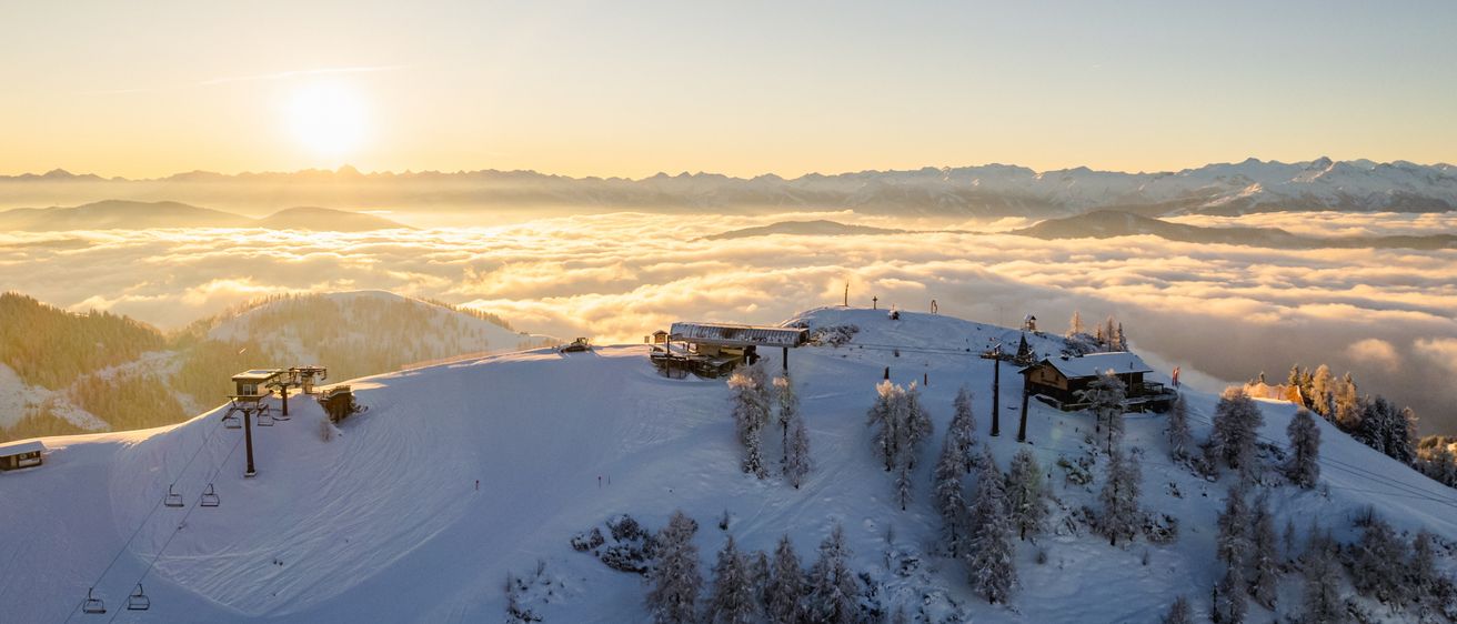 An aerial view of a snowy mountain landscape at sunset, with clouds below and a clear sky above. The ground is covered in snow, and there are ski lifts and buildings visible.